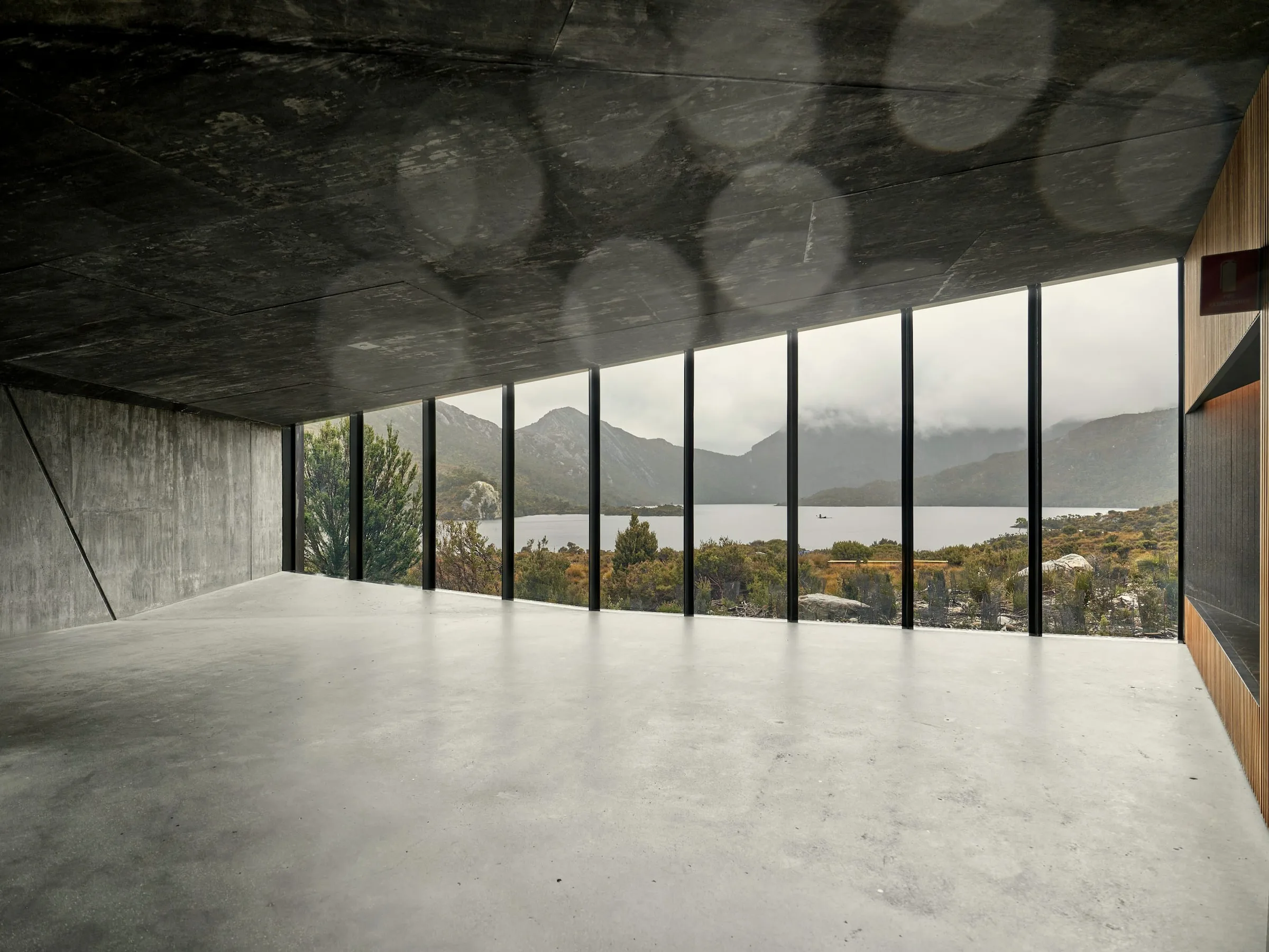 Concrete living space with a band of tall windows opening onto a lake and distant mountains